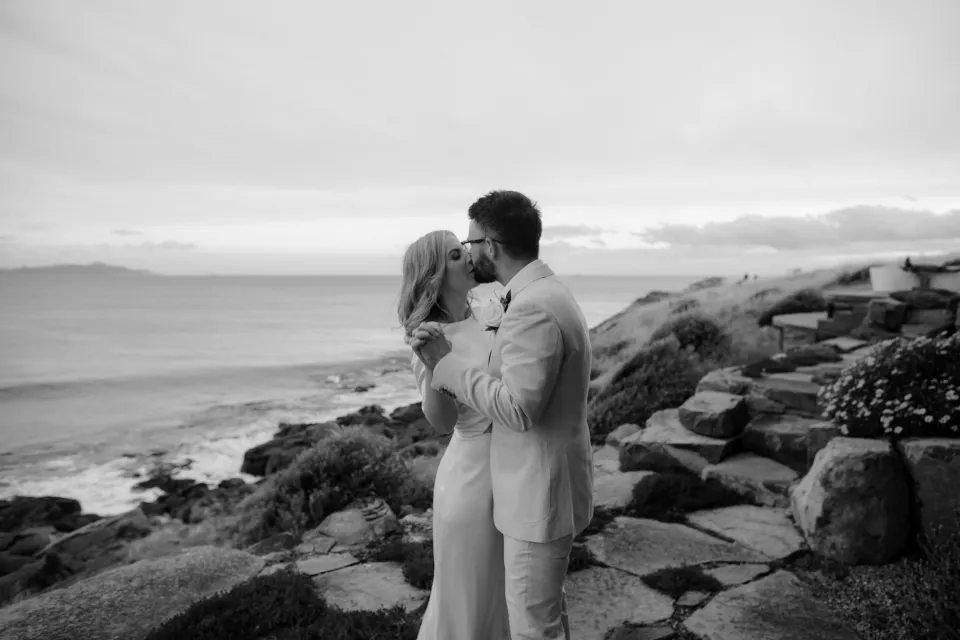 Black and white portrait of the couple kissing with Great Oyster Bay behind them, at Thalia Haven on the East Coast of Tasmania, Australia, for an Elopement Collective elopement. Photograph by Joey Willis.