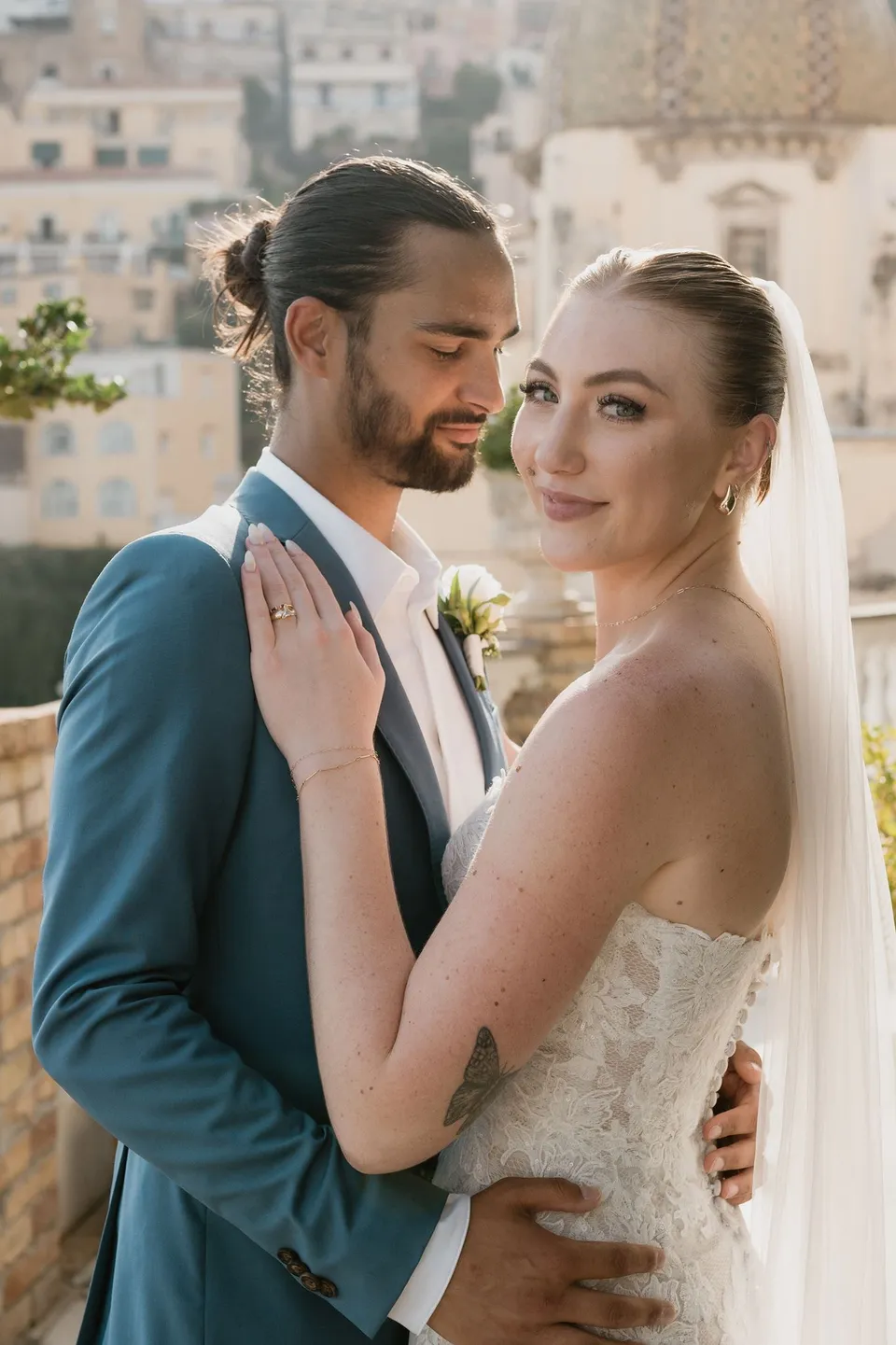 Close portrait of Skyler looking towards the camera as Zach leans towards her, with Positano’s hillside houses behind them, at Hotel Buca di Bacco in Positano, Amalfi Coast, Italy, for an Elopement Collective elopement. Photograph by Jason Corroto.