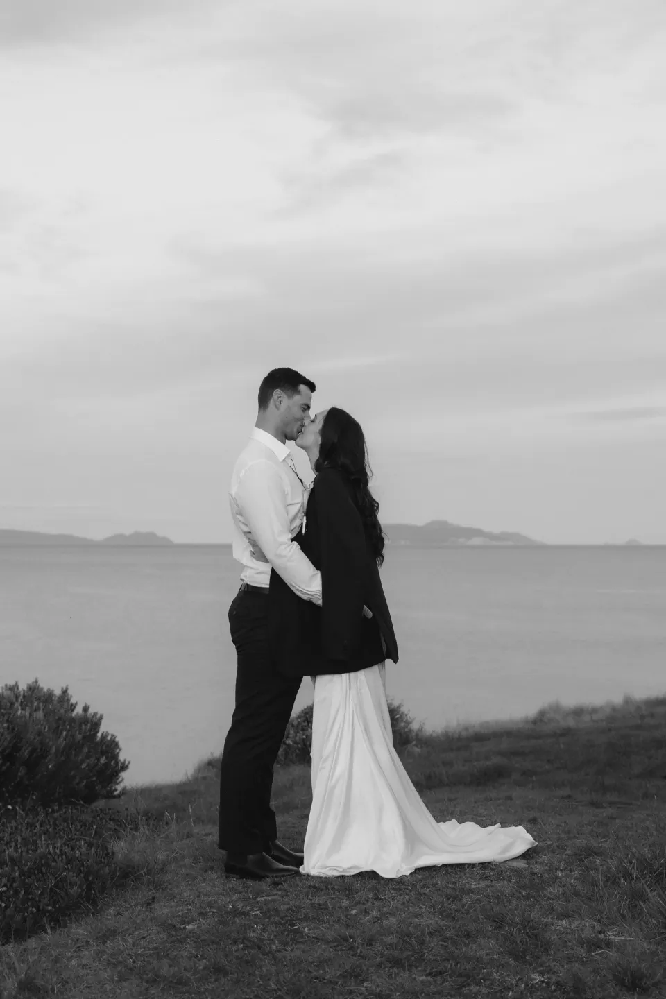 Black and white portrait of Dani and Anthony sharing a kiss above the bay, with his jacket around her shoulders, at Thalia Haven on Tasmania’s east coast, for an Elopement Collective elopement. Photograph by Bulb Creative.