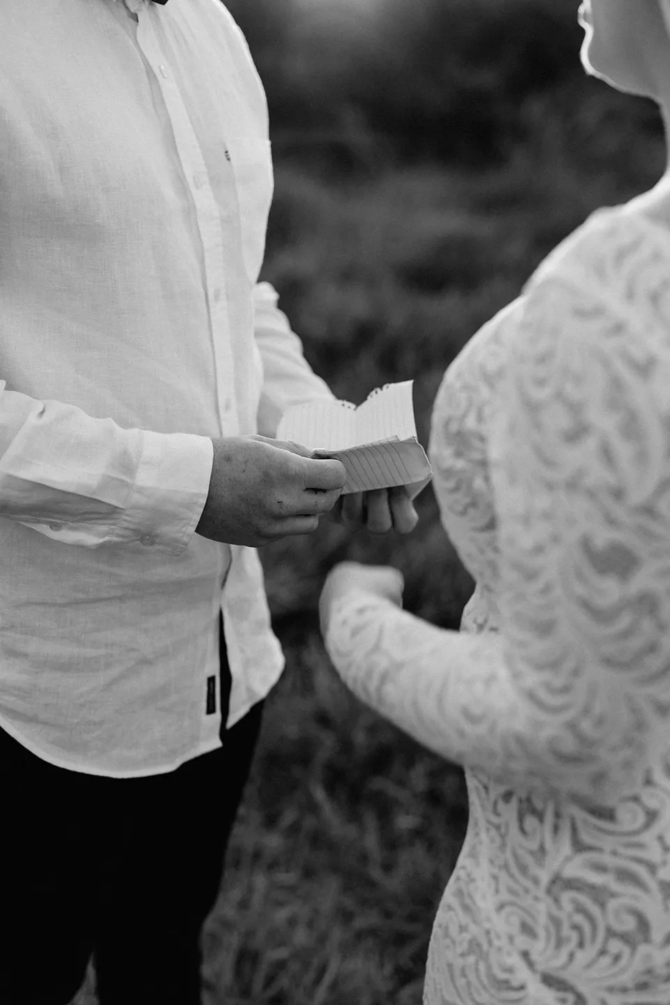 A black and white detail of Matt holding a small card beside Laura’s hand and lace sleeve during their ceremony, in the Tweed Valley, NSW, for an Elopement Collective elopement. Photo by Bulb Creative.