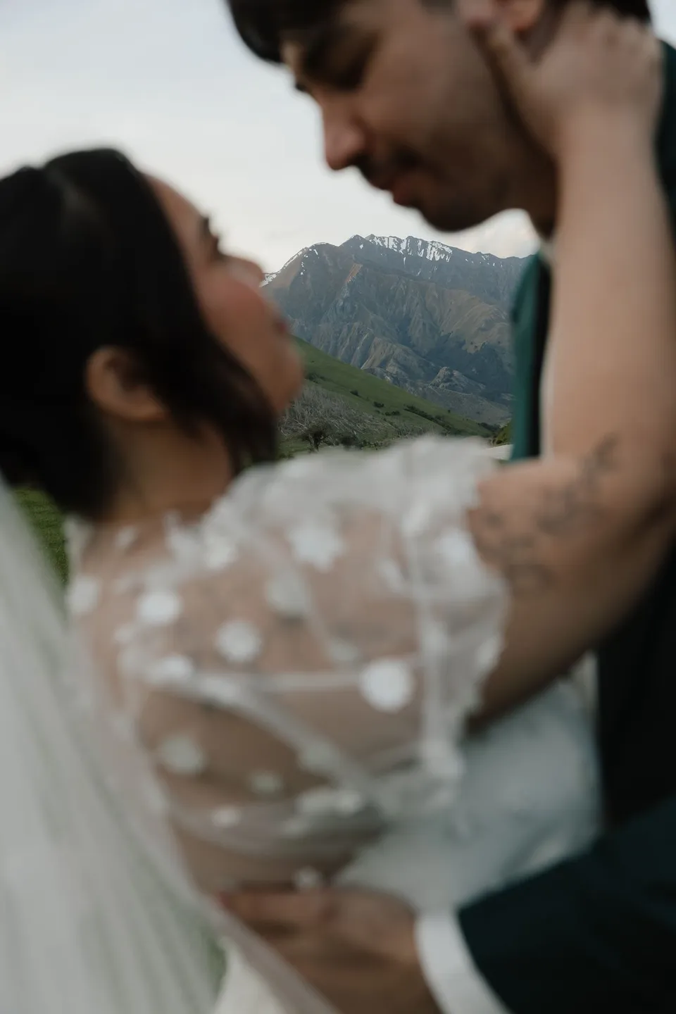 A close portrait of Adrian leaning towards Jaeda as they embrace, showing her sheer daisy-appliqué sleeves and his green suit, at Moke Lake, Queenstown, New Zealand, for an Elopement Collective Queenstown elopement. Photograph by Bulb Creative.
