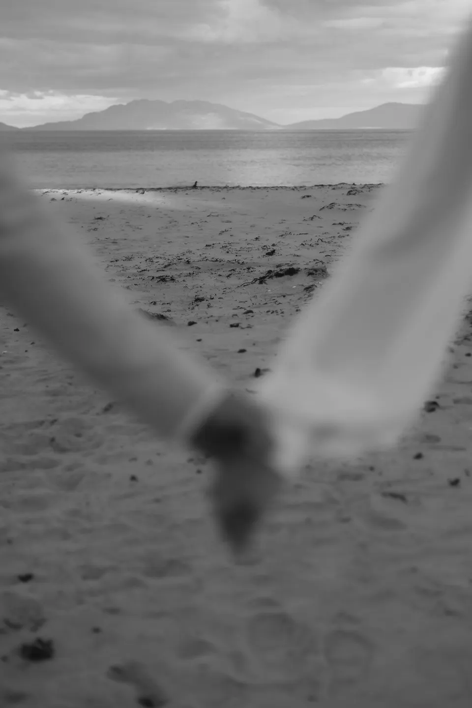 Black and white view of the bay and distant hills, framed by Renee and Mitch’s softly blurred joined hands. At Piermont Retreat on Tasmania’s east coast, for an Elopement Collective elopement. Photograph by Bulb Creative.
