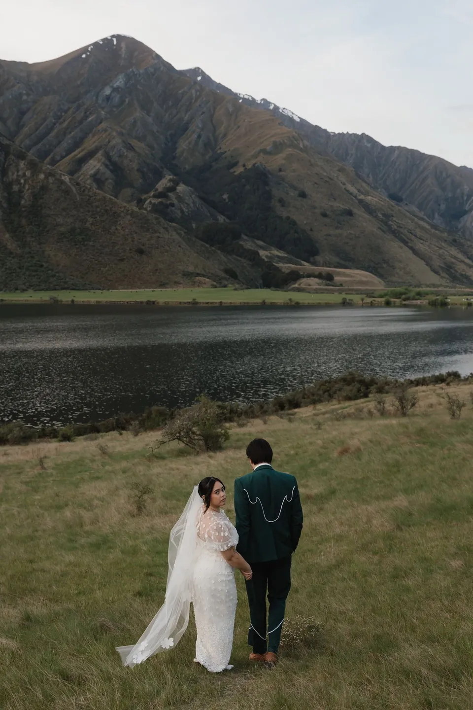 Jaeda and Adrian holding hands beside Moke Lake, with her veil trailing over the grass and steep hills across the water, at Moke Lake, Queenstown, New Zealand, for an Elopement Collective Queenstown elopement. Photograph by Bulb Creative.