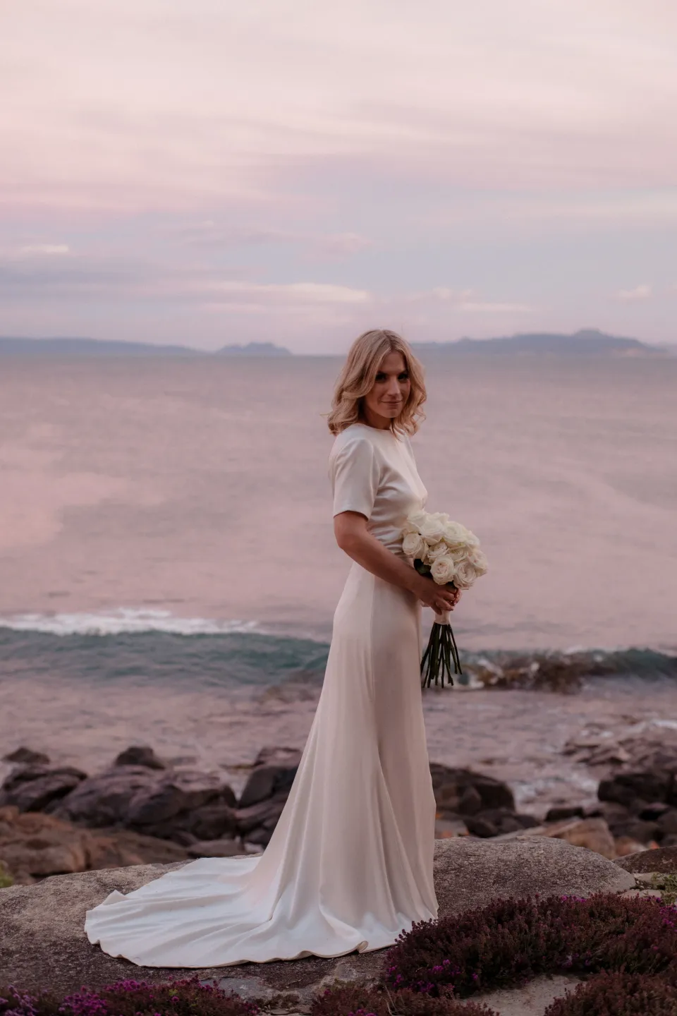 Jemmah turning towards the camera with her dress train beside the sea, at Thalia Haven on the East Coast of Tasmania, Australia, for an Elopement Collective elopement. Photograph by Joey Willis.