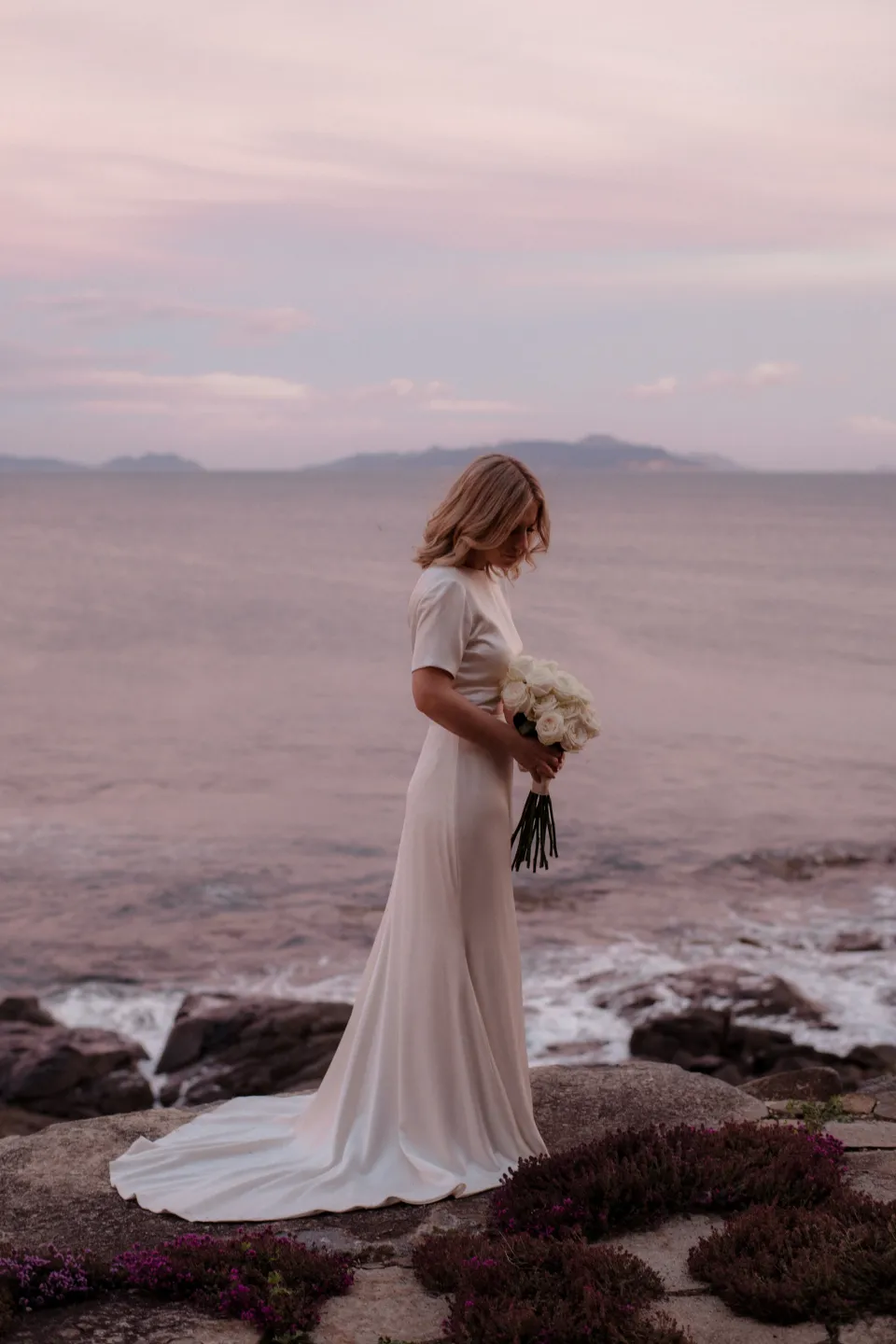 Jemmah standing on the terrace with her white bouquet by The Romantics in the evening light, at Thalia Haven on the East Coast of Tasmania, Australia, for an Elopement Collective elopement. Photograph by Joey Willis.