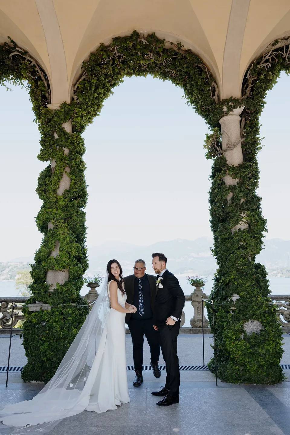 Lyndal and Lachlan standing with celebrant Josh Withers beneath Villa del Balbianello’s ivy-covered arches, with the lake behind them, at Lake Como in Lombardy, Italy, for an Elopement Collective elopement. Photograph by George Bowden.