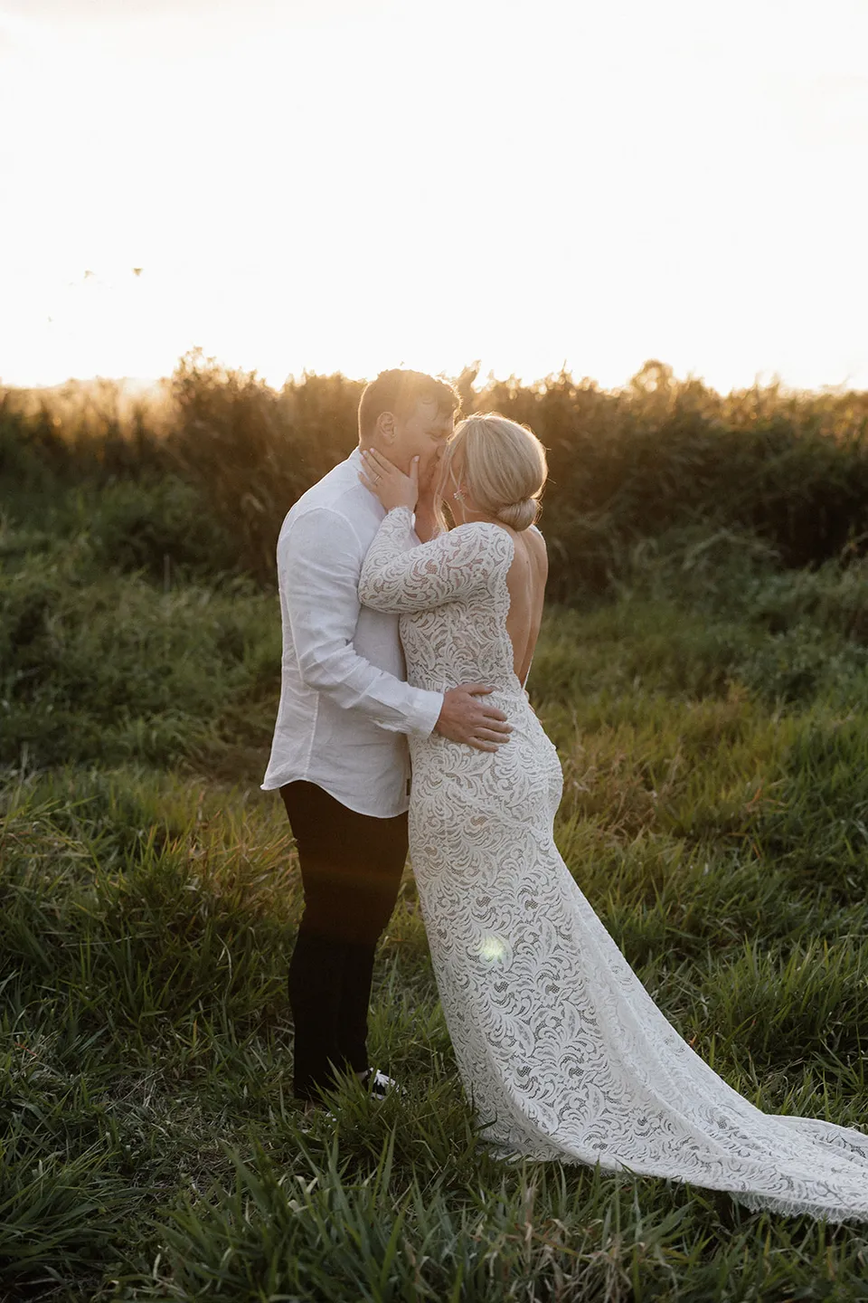 Matt and Laura kissing in the long grass after their ceremony, with Laura’s lace train spread behind her, in the Tweed Valley, NSW, for an Elopement Collective elopement. Photo by Bulb Creative.