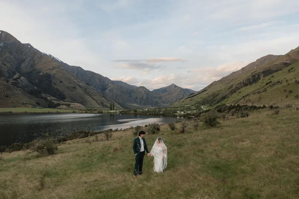 A wide view of Jaeda and Adrian beside Moke Lake, surrounded by green hills beneath a softly coloured evening sky, at Moke Lake, Queenstown, New Zealand, for an Elopement Collective Queenstown elopement. Photograph by Bulb Creative.