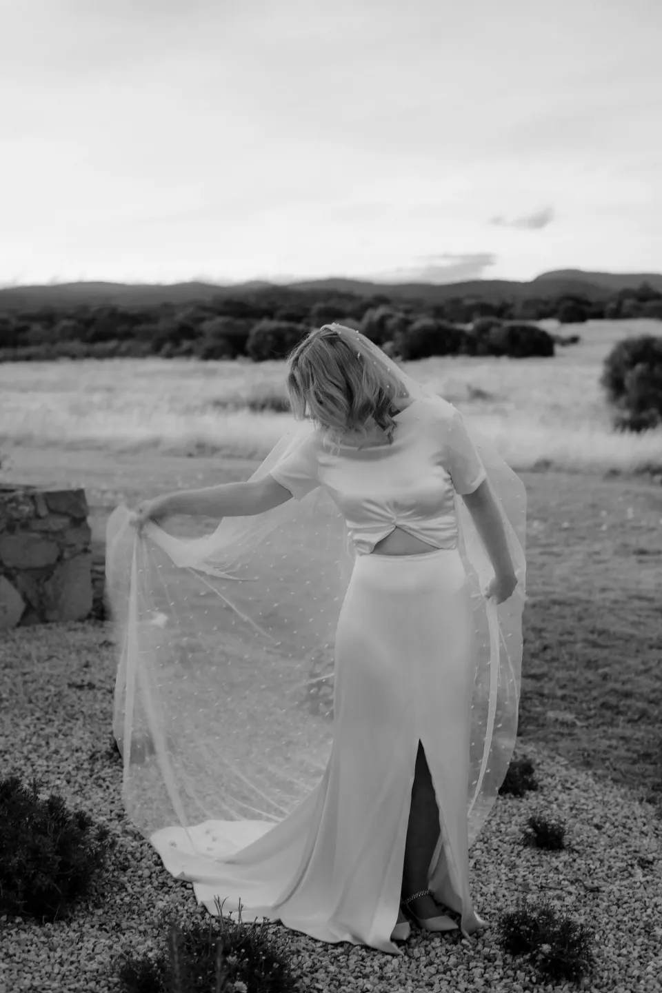 Black and white photograph of Jemmah moving her dress through the meadow, at Thalia Haven on the East Coast of Tasmania, Australia, for an Elopement Collective elopement. Photograph by Joey Willis.
