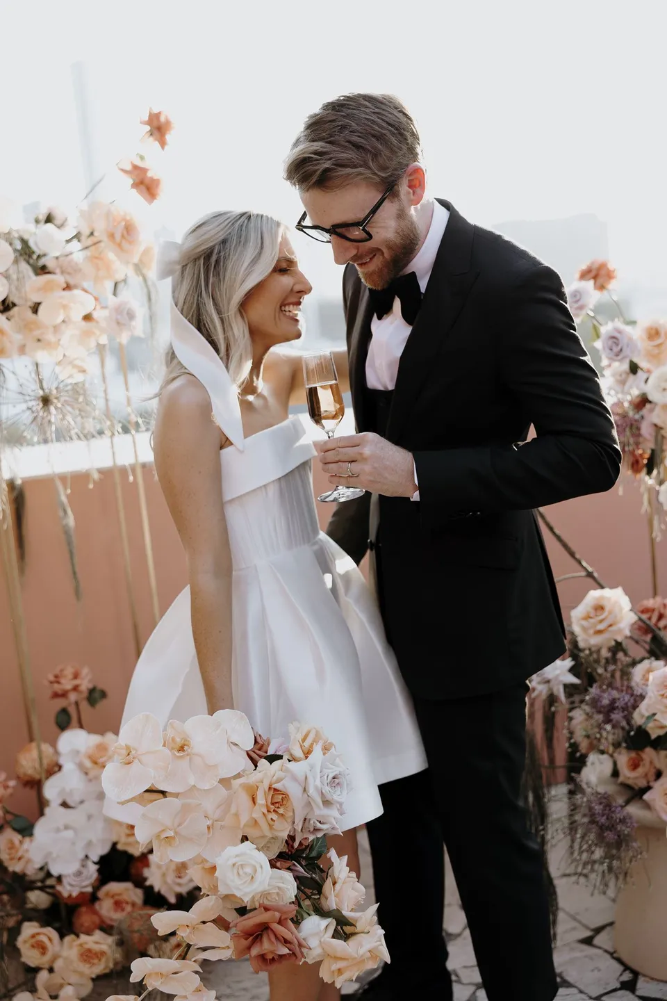 Danielle and Adam smiling together with champagne beside the terrace flowers by Mrs Gibbons Florals, at The Calile Hotel in Fortitude Valley, Brisbane, for an Elopement Collective elopement. Photo by George Bowden.