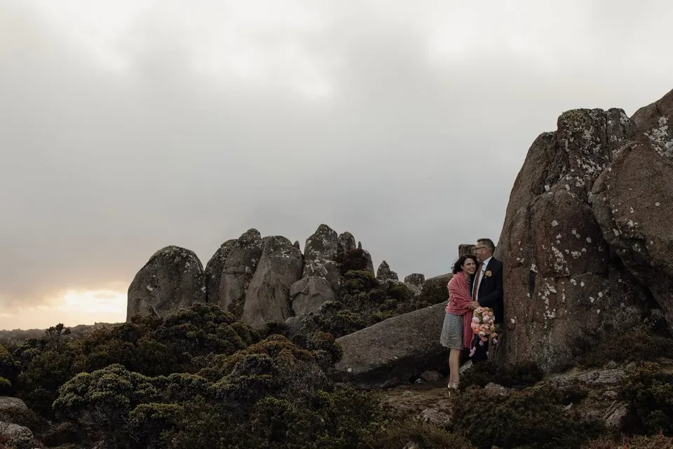 A couple stand among tall, lichen-covered boulders on a cloudy mountain plateau.