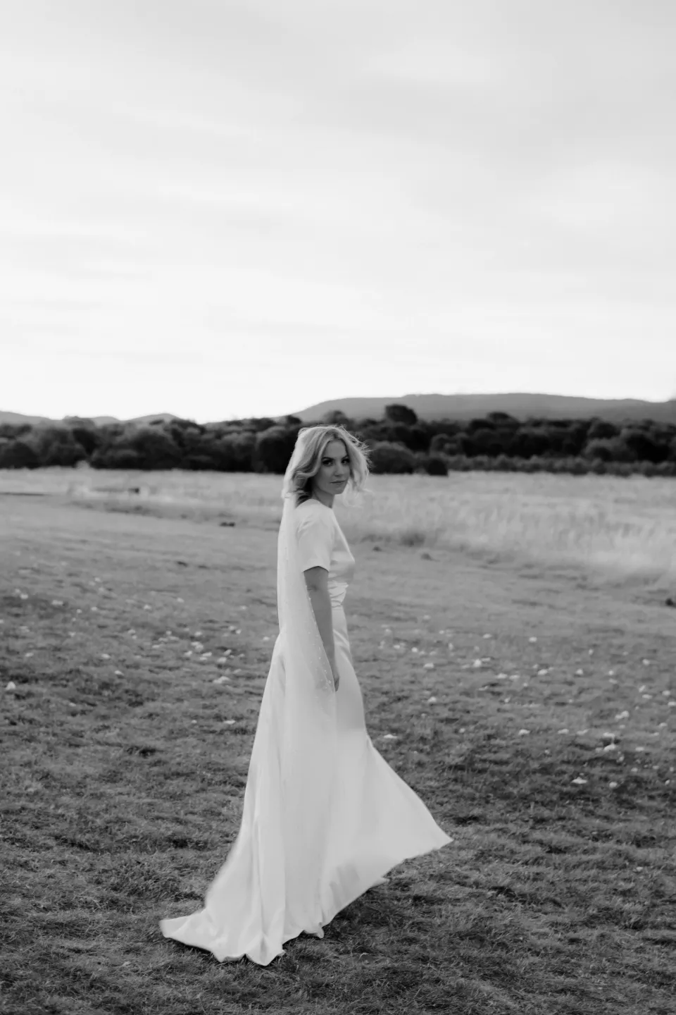 Black and white portrait of Jemmah looking back across the meadow, at Thalia Haven on the East Coast of Tasmania, Australia, for an Elopement Collective elopement. Photograph by Joey Willis.
