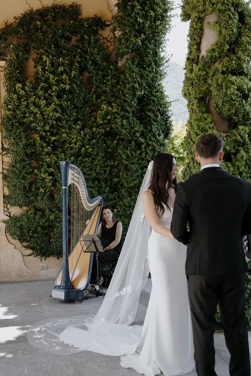 Lyndal and Lachlan standing together beside a harpist playing beneath Villa del Balbianello’s ivy-covered loggia, at Lake Como in Lombardy, Italy, for an Elopement Collective elopement. Photograph by George Bowden.