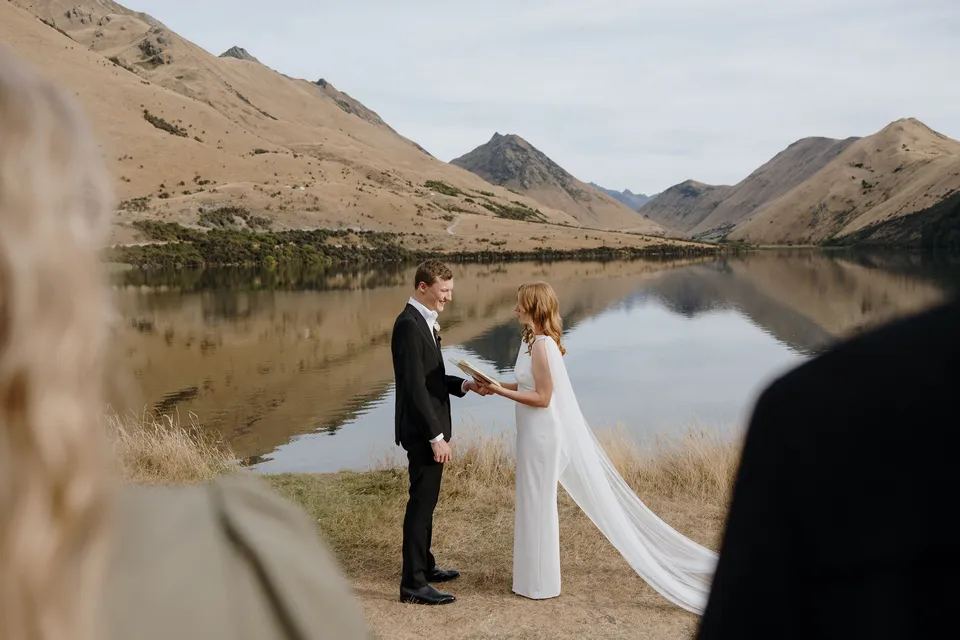 Neve and Hamish facing each other during their vows, framed by guests and mountains reflected in the lake, at Moke Lake in Queenstown, New Zealand, for an Elopement Collective Queenstown elopement. Photo by George Bowden.