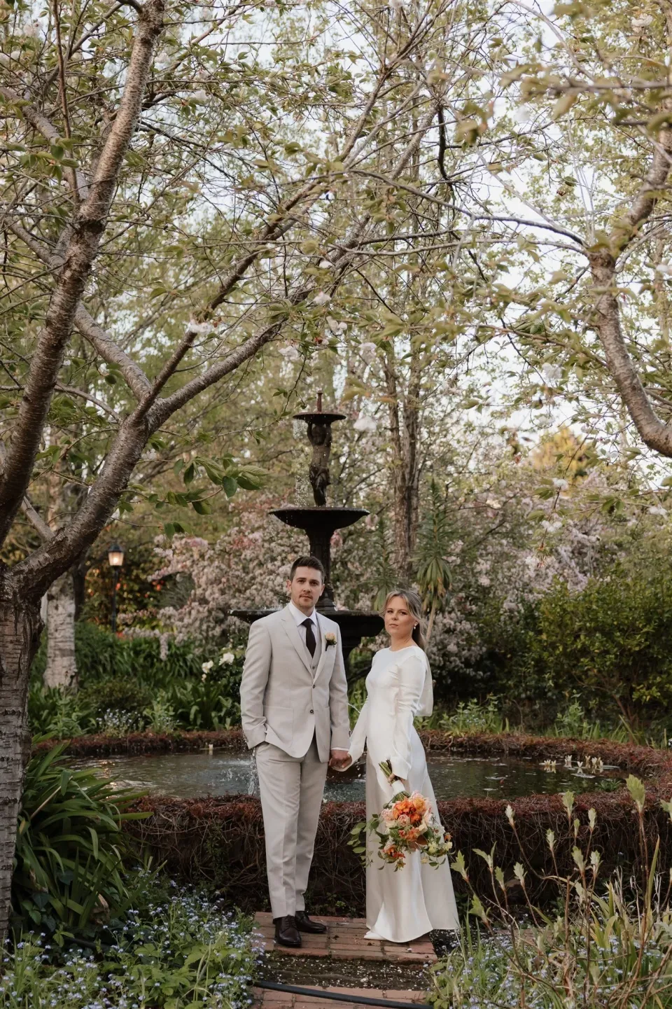 Maddy and Cameron standing hand in hand on the garden path with her bouquet by The Flower Jar and a fountain behind them, at The Mayor’s Residence in Daylesford, Victoria, for an Elopement Collective elopement. Photo by Bulb Creative.