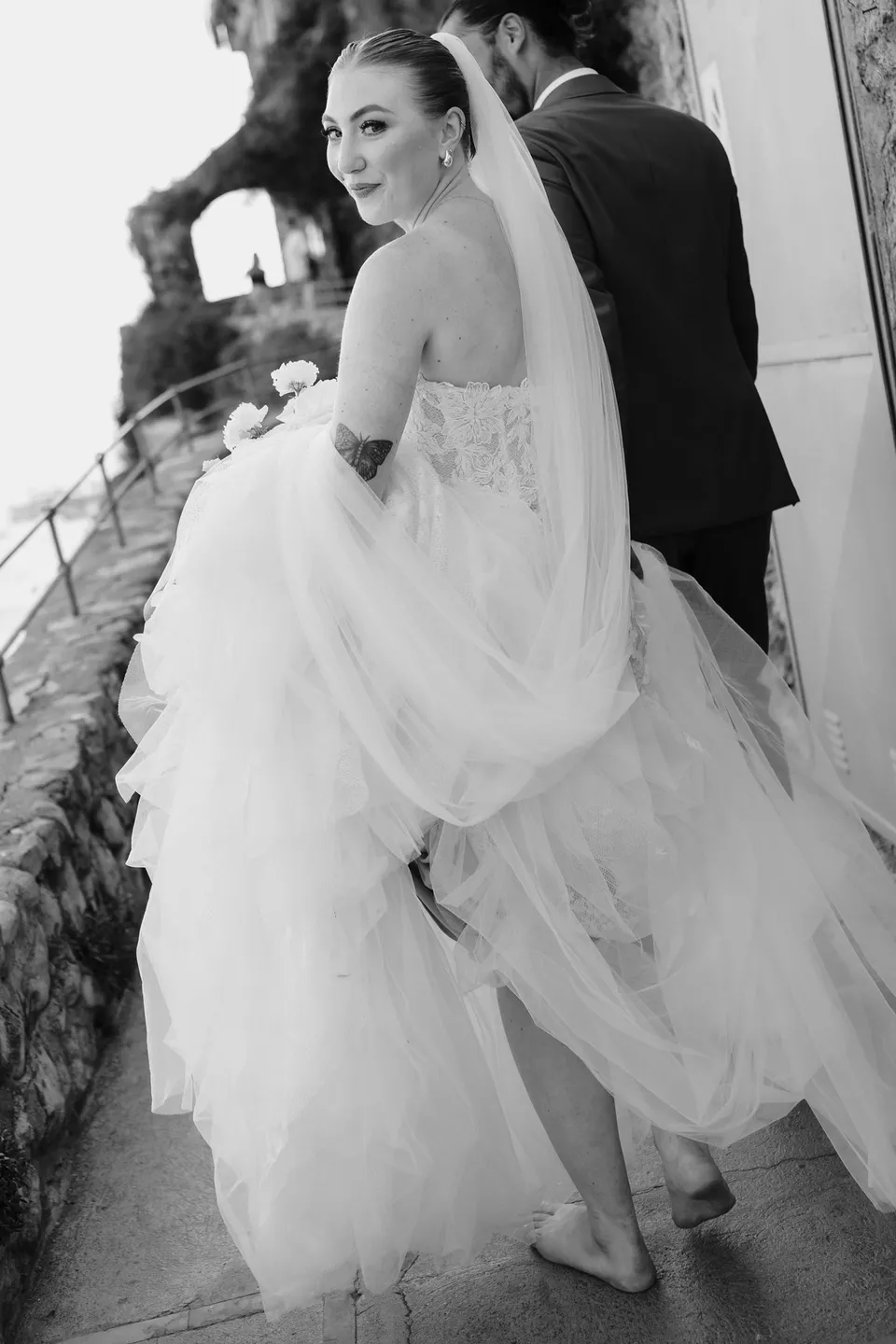 Black and white photograph of Skyler looking back over her shoulder while holding her dress and following Zach down the steps, in Positano on the Amalfi Coast, Italy, for an Elopement Collective elopement. Photograph by Jason Corroto.