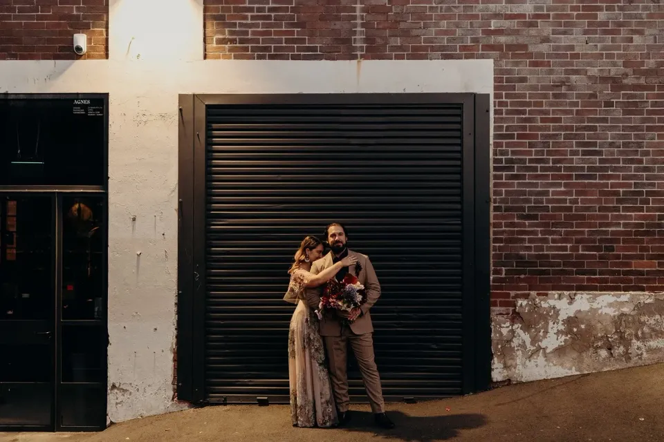 Wide evening view of Victoria and Daniel embracing against a brick building and black roller door, in Fortitude Valley, Brisbane, for an Elopement Collective elopement. Photo by Tessa Shannon.