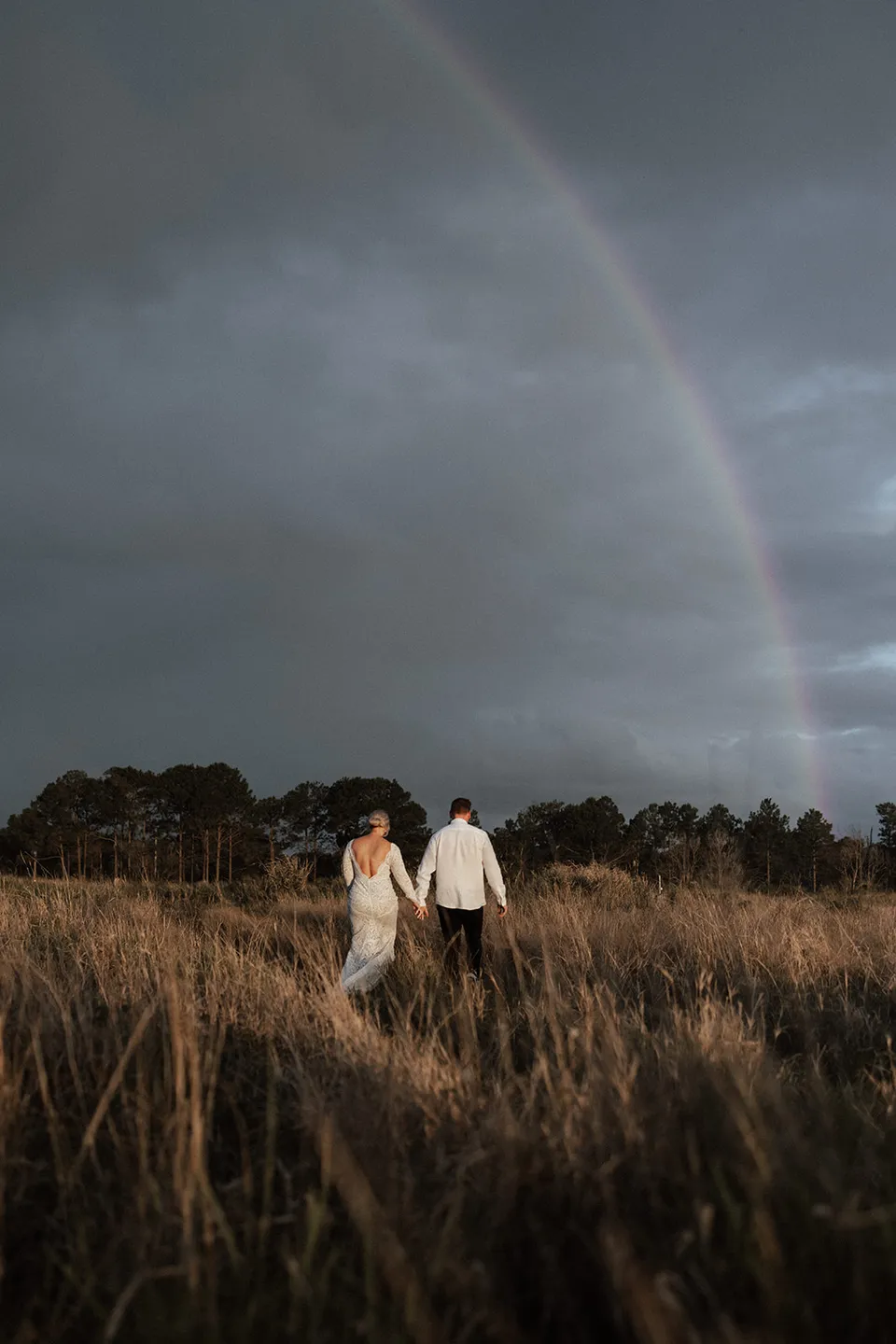 Matt and Laura walking hand in hand through long grass beneath a rainbow and grey skies, in the Tweed Valley, NSW, for an Elopement Collective elopement. Photo by Bulb Creative.