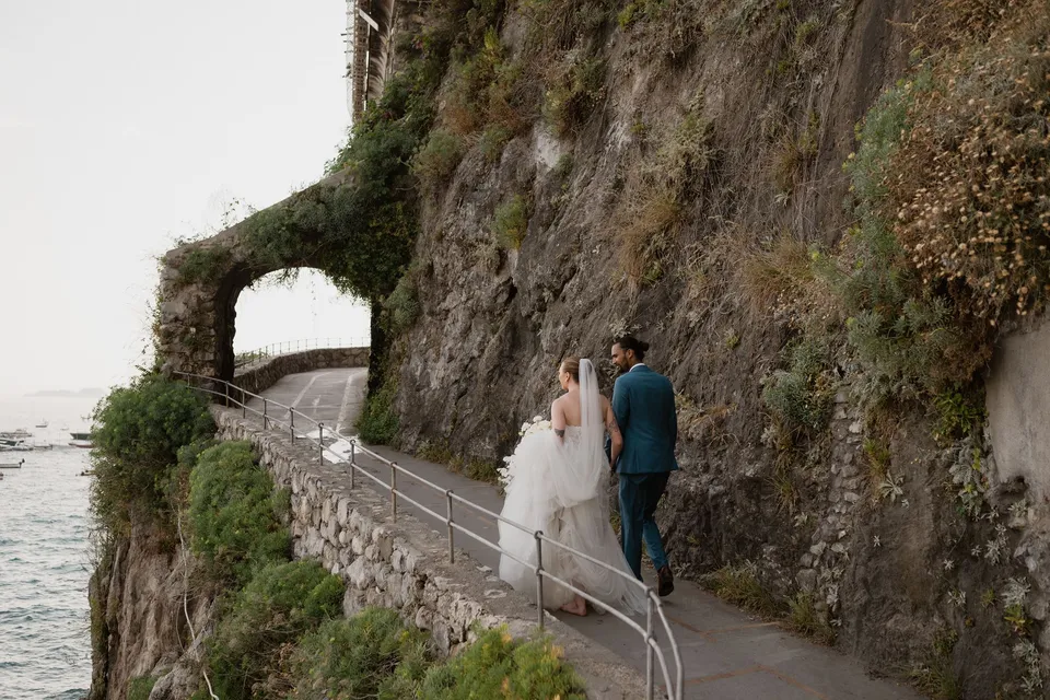 Skyler and Zach walking together along a cliffside path beneath an ivy-covered arch beside a stone wall, in Positano on the Amalfi Coast, Italy, for an Elopement Collective elopement. Photograph by Jason Corroto.