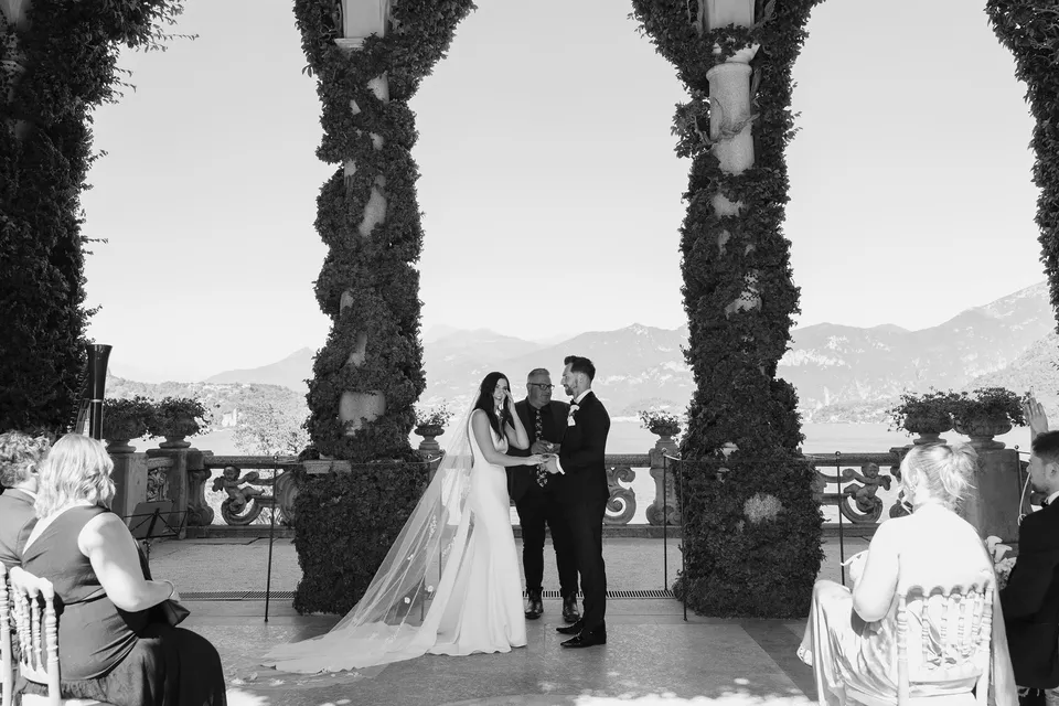 Black and white wide view of Lyndal and Lachlan’s ceremony with Josh Withers, a small group of seated guests and mountains beyond Villa del Balbianello’s loggia, at Lake Como in Lombardy, Italy, for an Elopement Collective elopement. Photograph by George Bowden.