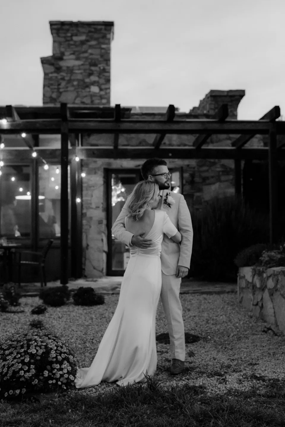 Black and white photograph of Jemmah and Jack embracing outside the illuminated stone house, at Thalia Haven on the East Coast of Tasmania, Australia, for an Elopement Collective elopement. Photograph by Joey Willis.