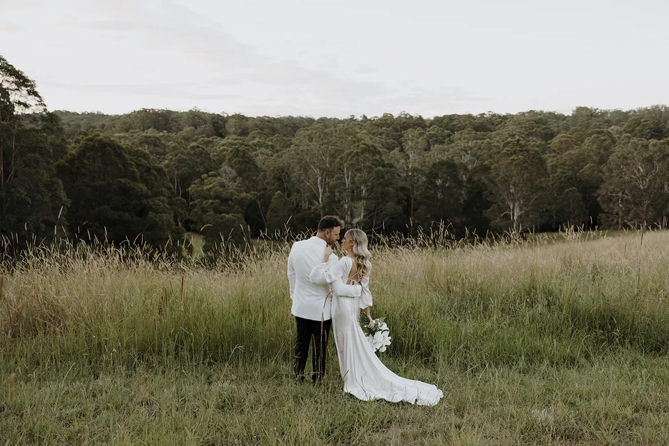 Gabrielle and Greg embracing in tall grass with a wooded ridge behind them, at Roundhill Retreat in Obi Obi, Sunshine Coast Hinterland, Queensland, for an Elopement Collective elopement. Photo by George Bowden.
