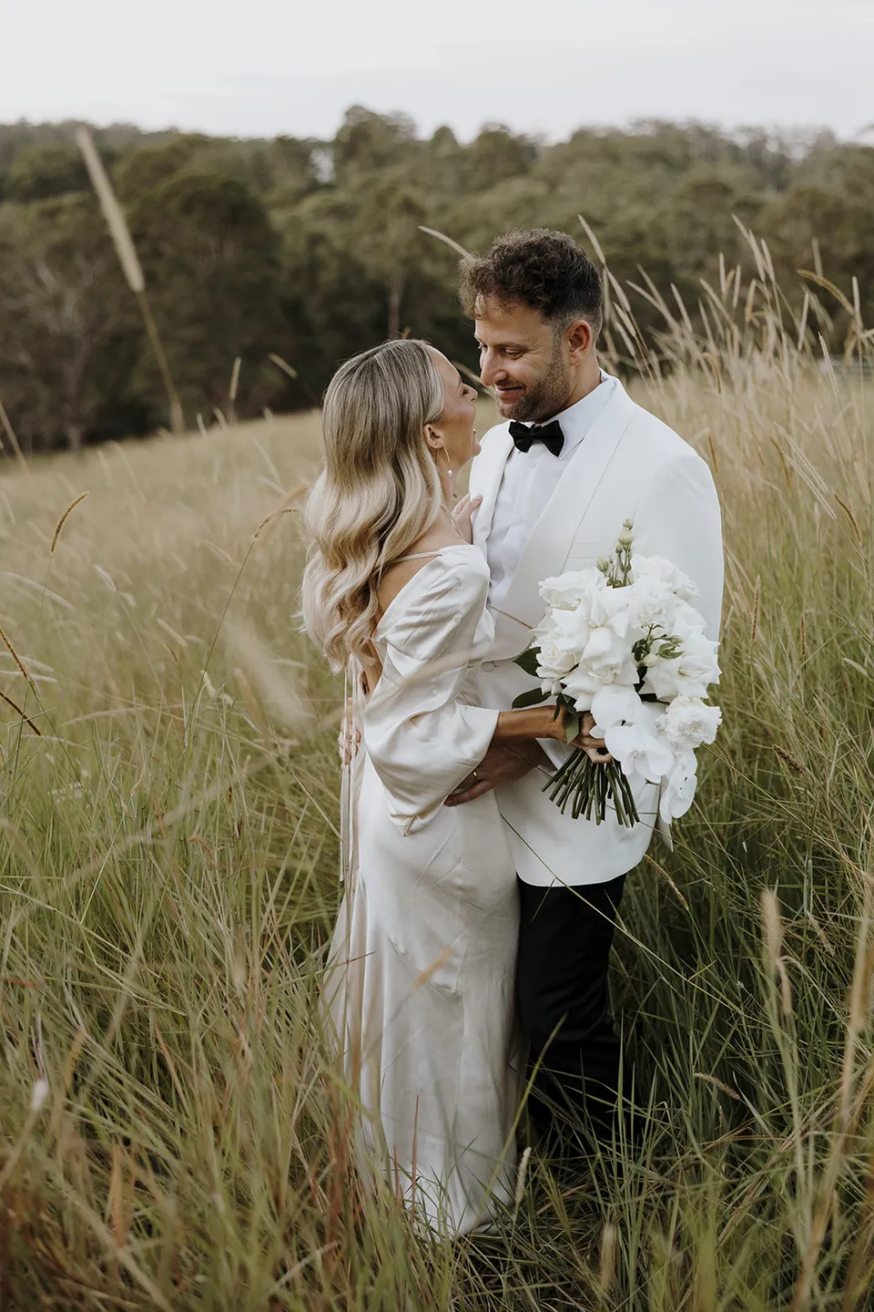Gabrielle holding her white bouquet by Mrs Gibbons as Greg smiles at her among the tall paddock grasses, at Roundhill Retreat in Obi Obi, Sunshine Coast Hinterland, Queensland, for an Elopement Collective elopement. Photo by George Bowden.