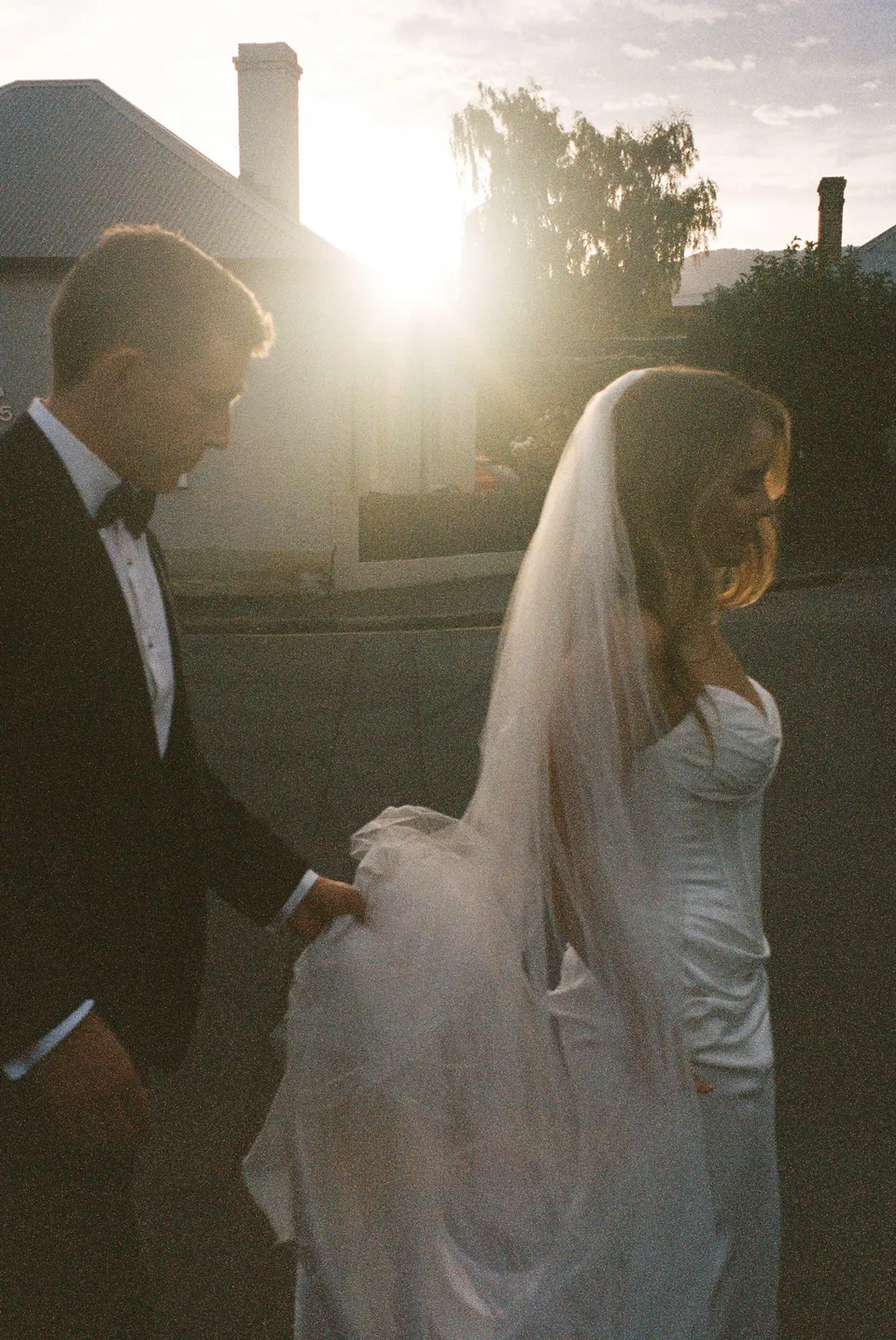 Tanesha and Andrew holding hands as low sunlight streams between the cottages behind them, at Battery Point in Hobart, Tasmania, Australia, for an Elopement Collective summer elopement. Photograph by George Bowden.