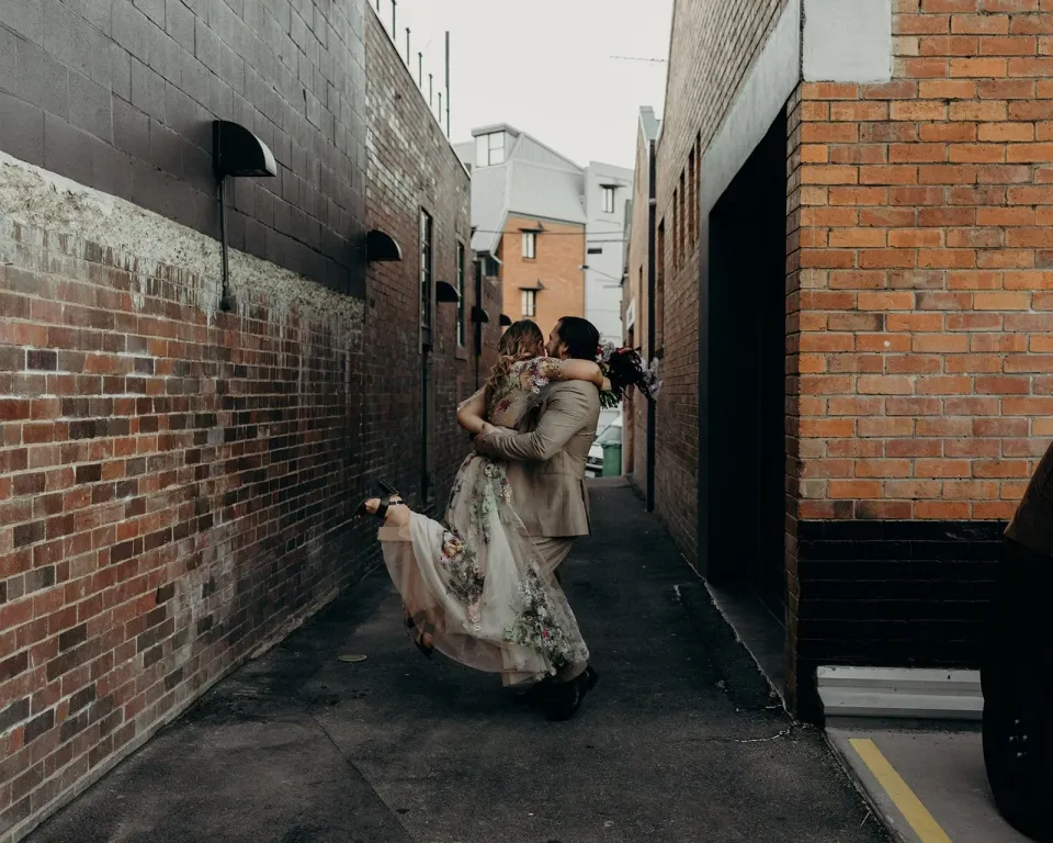 Daniel lifting Victoria into an embrace in a narrow brick-lined laneway, in Fortitude Valley, Brisbane, for an Elopement Collective elopement. Photo by Tessa Shannon.