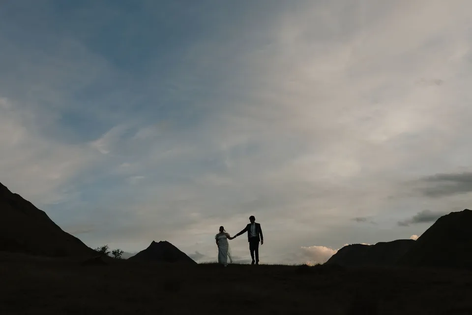 Jaeda and Adrian silhouetted against the evening sky as they hold hands between the mountain ridgelines, at Moke Lake, Queenstown, New Zealand, for an Elopement Collective Queenstown elopement. Photograph by Bulb Creative.