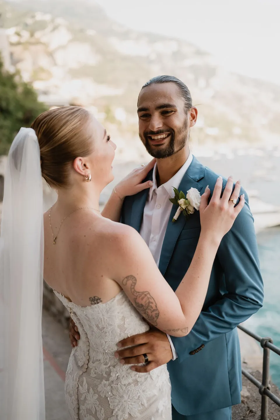 Zach laughing as Skyler rests a hand on his chest, with his white buttonhole by Flora Garden - Positano visible on his blue suit, in Positano on the Amalfi Coast, Italy, for an Elopement Collective elopement. Photograph by Jason Corroto.