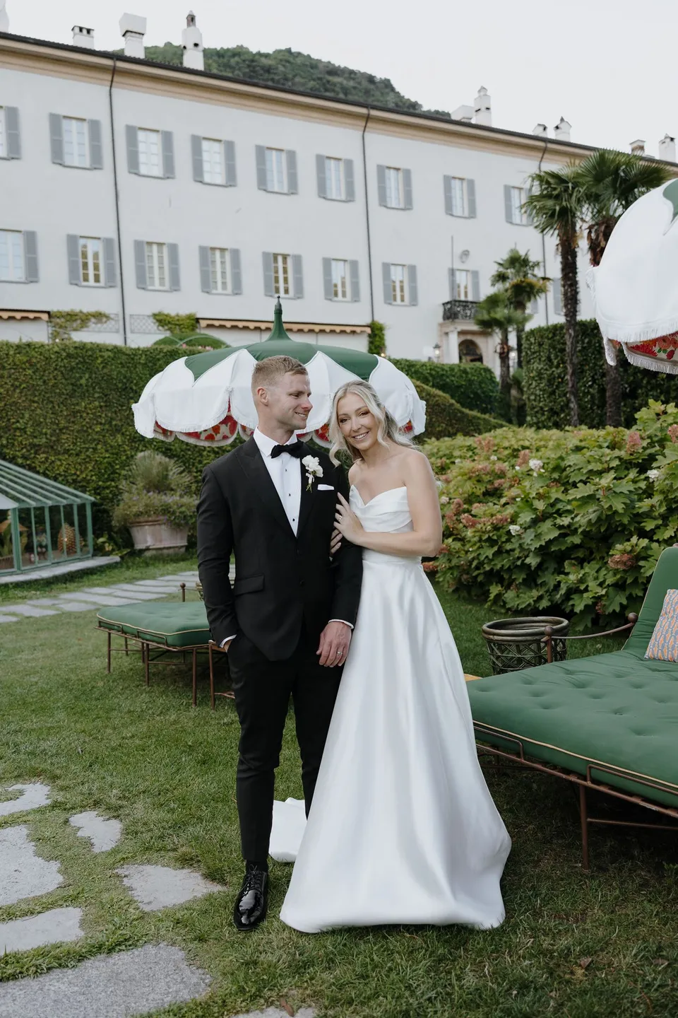 Zoe and Charlton facing one another beside green loungers and scalloped white parasols, with the villa behind them, at Passalacqua in Moltrasio, Lake Como, Italy, for an Elopement Collective elopement. Photo by George Bowden.