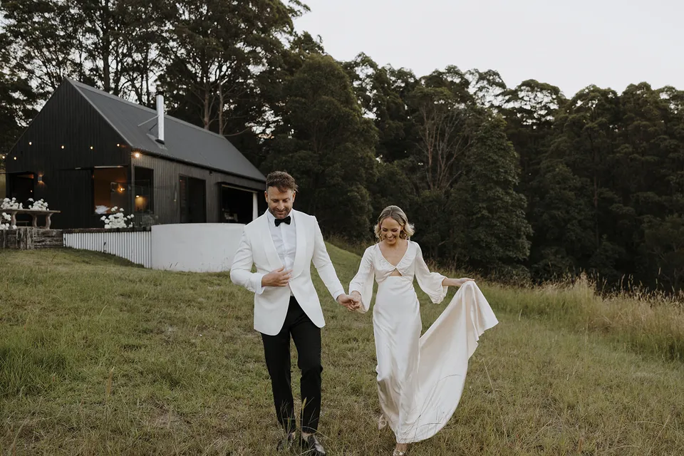 Gabrielle and Greg walking hand in hand through the paddock with the dark cabin and festoon lights behind them, at Roundhill Retreat in Obi Obi, Sunshine Coast Hinterland, Queensland, for an Elopement Collective elopement. Photo by George Bowden.