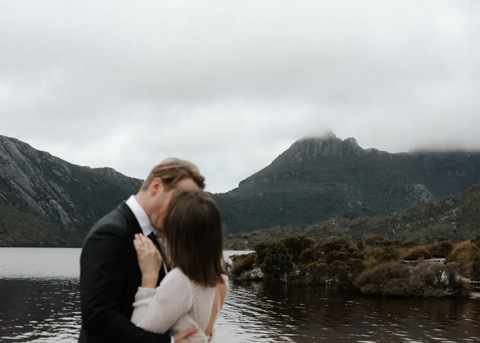 April and Josh kissing with mist drifting around the mountain peaks behind them, at Dove Lake, Cradle Mountain, Tasmania, for an Elopement Collective elopement. Photo by Bulb Creative.