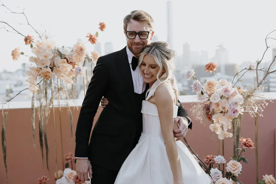 Adam embracing Danielle from behind as she smiles among the terrace flower arrangements by Mrs Gibbons Florals, at The Calile Hotel in Fortitude Valley, Brisbane, for an Elopement Collective elopement. Photo by George Bowden.
