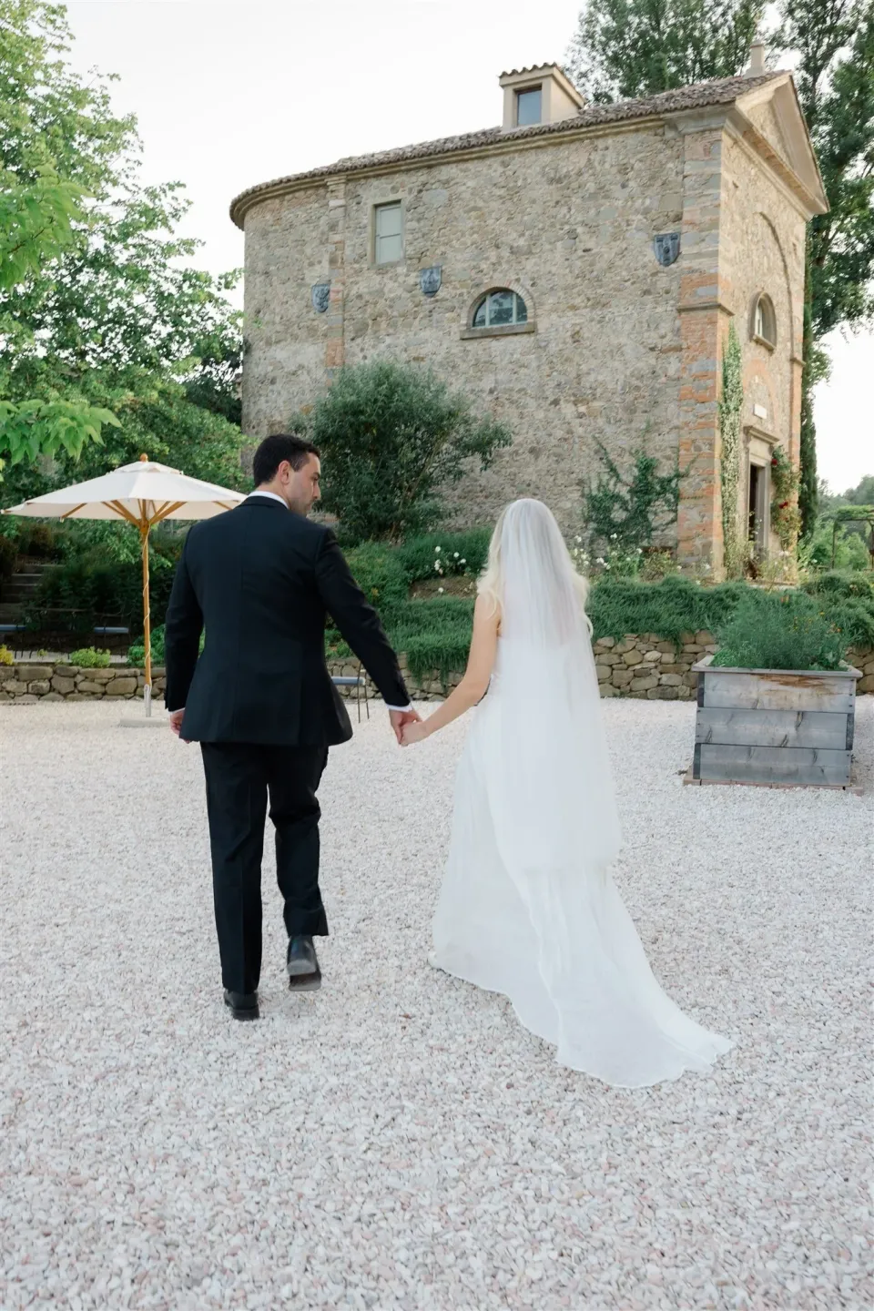 Shelby and Jack walking hand in hand across a gravel courtyard, her veil and dress trailing behind her, in Umbria, Italy, for an Elopement Collective elopement. Photo by James from House of Love.