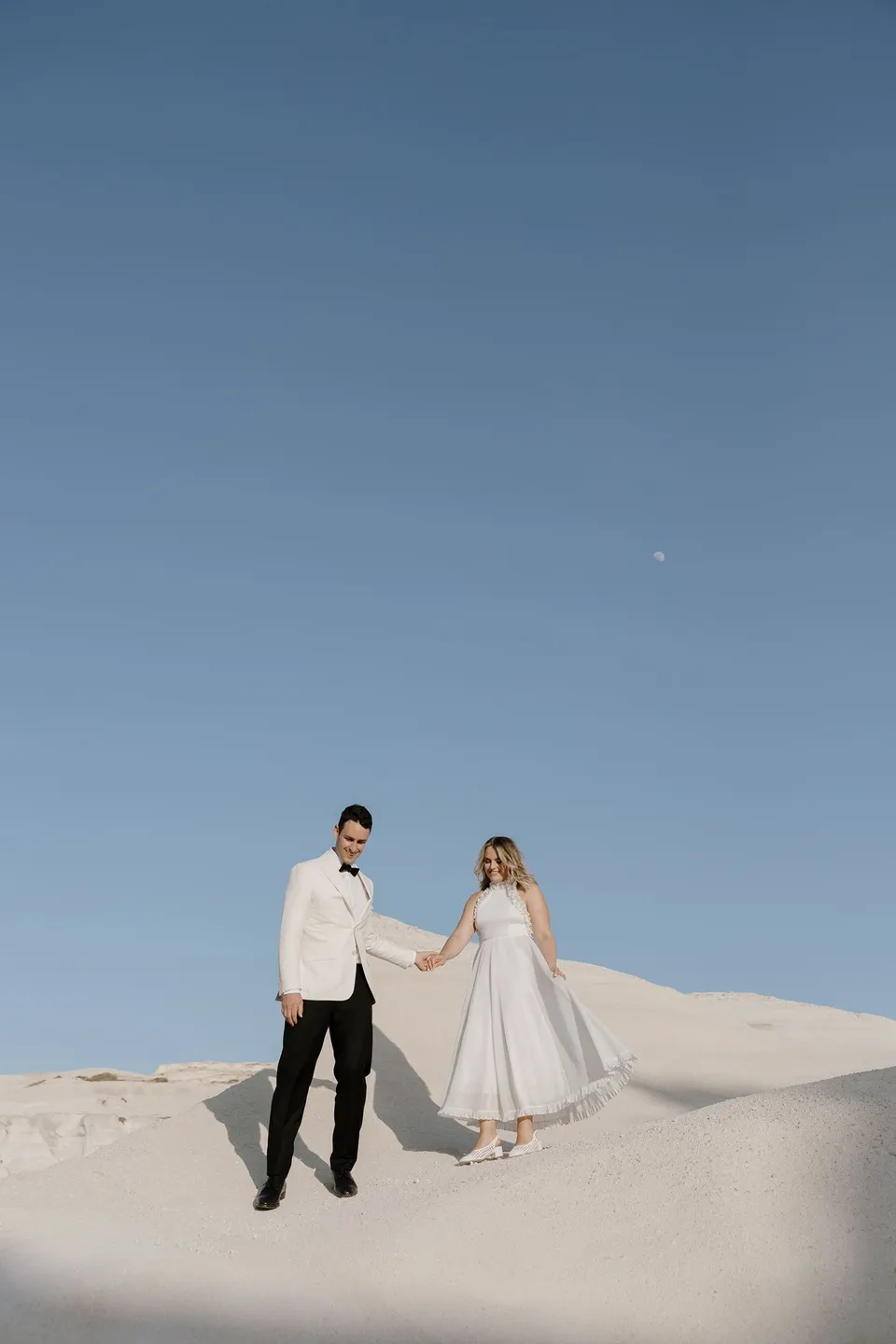 Georgina and Brad holding hands on a white rock ridge beneath a broad blue sky and a small visible moon, at Sarakiniko on Milos in the Cyclades, Greece, during an Elopement Collective elopement. Photograph by George Bowden.