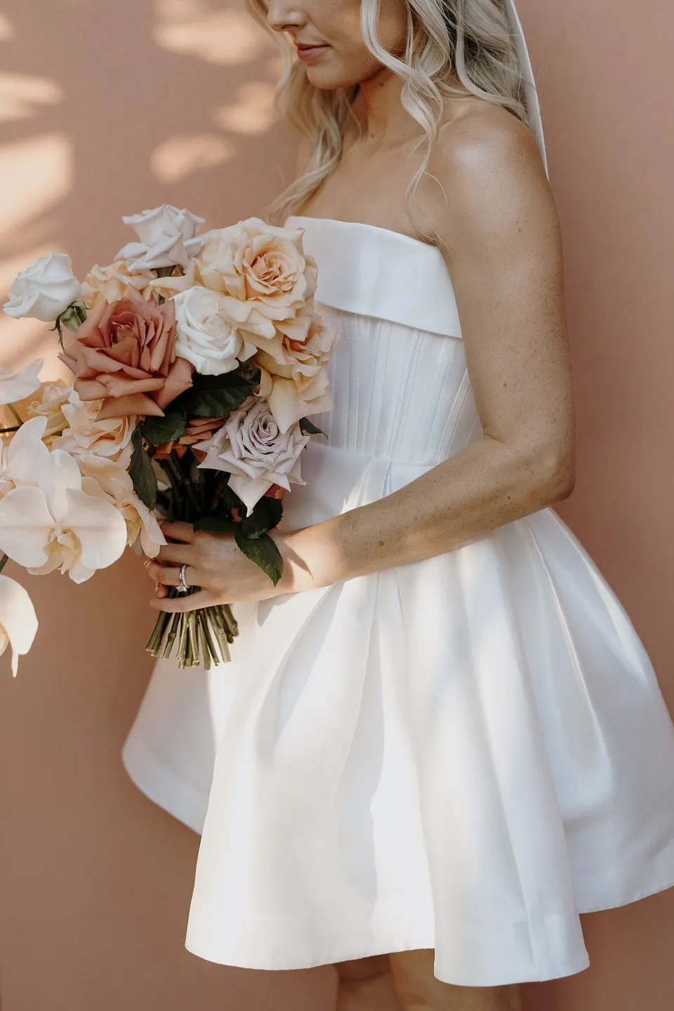 Close view of Danielle’s structured white dress and bouquet of peach roses and pale orchids by Mrs Gibbons Florals, at The Calile Hotel in Fortitude Valley, Brisbane, for an Elopement Collective elopement. Photo by George Bowden.