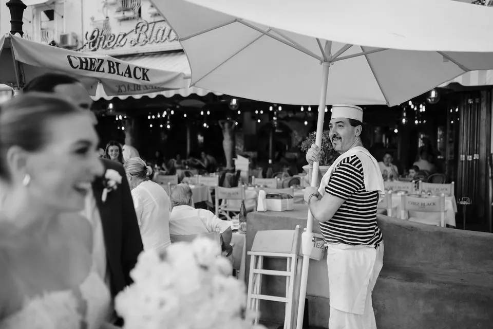 Black and white photograph of Skyler and Zach passing smiling restaurant staff beside outdoor tables and parasols, in Positano on the Amalfi Coast, Italy, for an Elopement Collective elopement. Photograph by Jason Corroto.