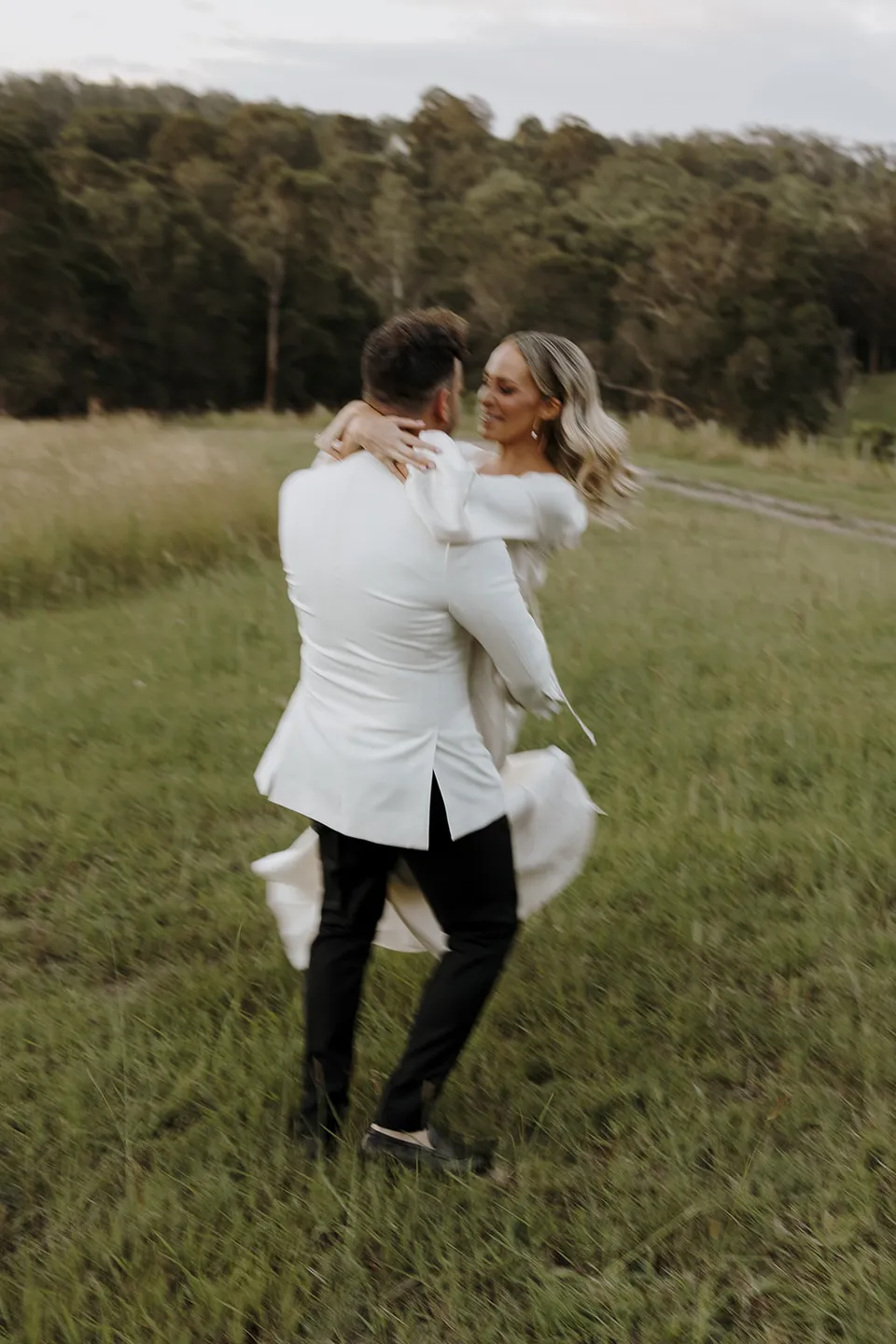 Greg carrying Gabrielle through the grassy paddock as she wraps her arms around his shoulders, at Roundhill Retreat in Obi Obi, Sunshine Coast Hinterland, Queensland, for an Elopement Collective elopement. Photo by George Bowden.