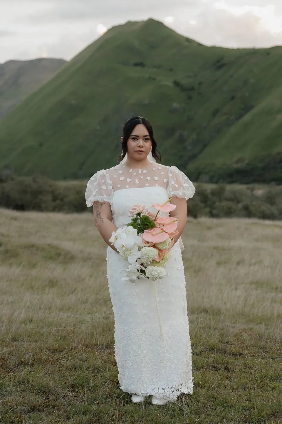 Jaeda holding her pink and white Orange Blossom Designs bouquet in front of the green hills, at Moke Lake, Queenstown, New Zealand, for an Elopement Collective Queenstown elopement. Photograph by Bulb Creative.