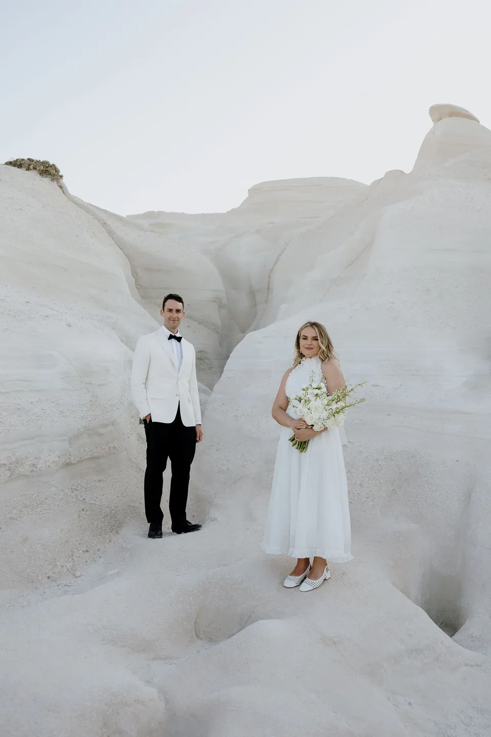 Georgina and Brad standing together in a curving white rock gully, with her white bouquet by Giasemi, at Sarakiniko on Milos in the Cyclades, Greece, during an Elopement Collective elopement. Photograph by George Bowden.