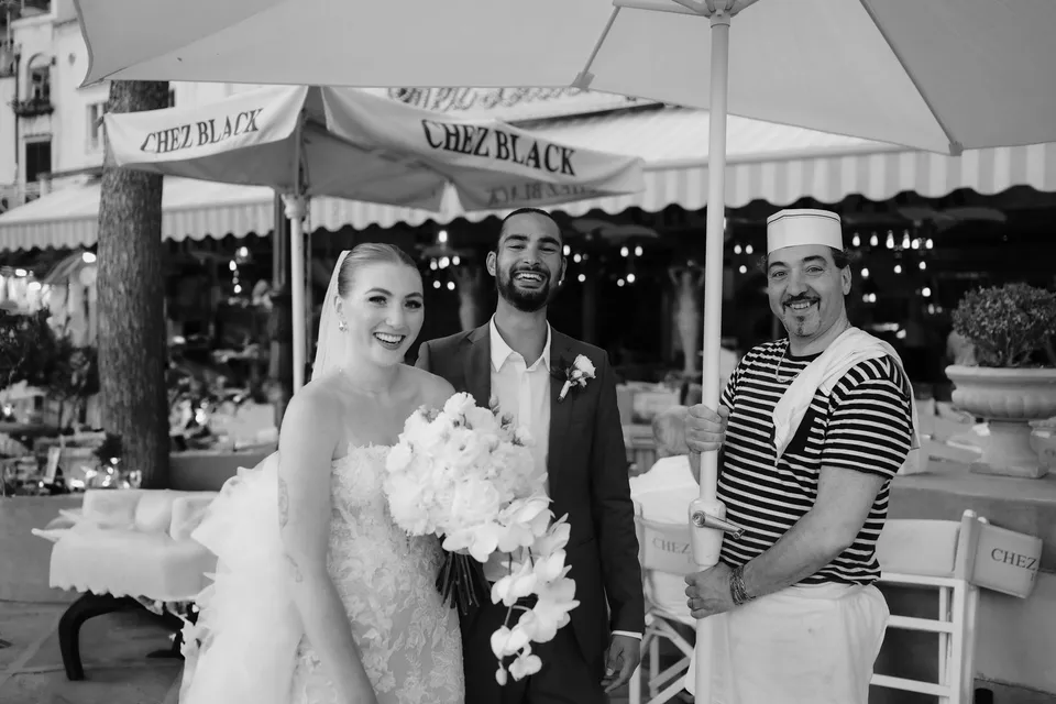 Black and white portrait of Skyler and Zach smiling with a restaurant staff member beneath a parasol, with her Flora Garden - Positano bouquet, in Positano on the Amalfi Coast, Italy, for an Elopement Collective elopement. Photograph by Jason Corroto.