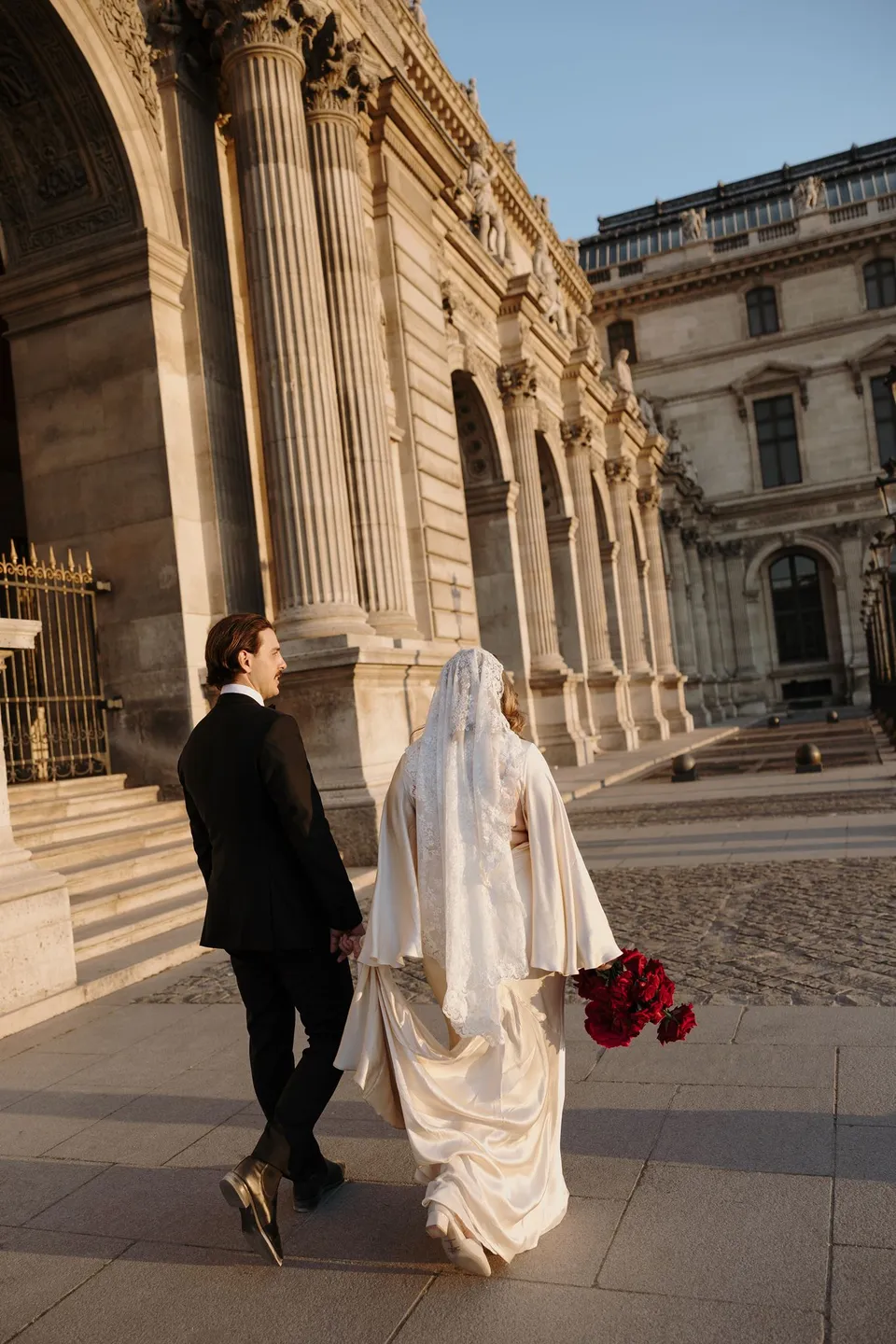 Rear view of Kara and Samuel walking hand in hand across the Louvre forecourt, with her red bouquet and veil trailing behind, at the Louvre in Paris, France, during an Elopement Collective elopement. Photograph by George Bowden.