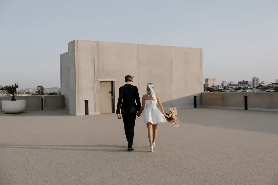 Danielle and Adam walking away hand in hand across the open hotel rooftop, at The Calile Hotel in Fortitude Valley, Brisbane, for an Elopement Collective elopement. Photo by George Bowden.