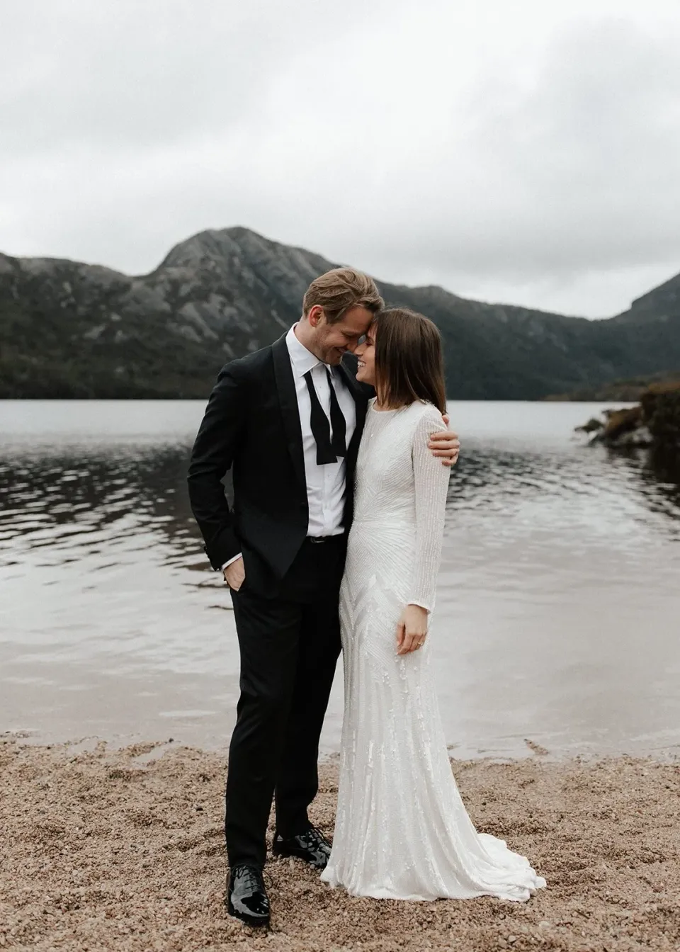 April and Josh touching foreheads on the sandy lakeshore, her dress trailing beside them, at Dove Lake, Cradle Mountain, Tasmania, for an Elopement Collective elopement. Photo by Bulb Creative.