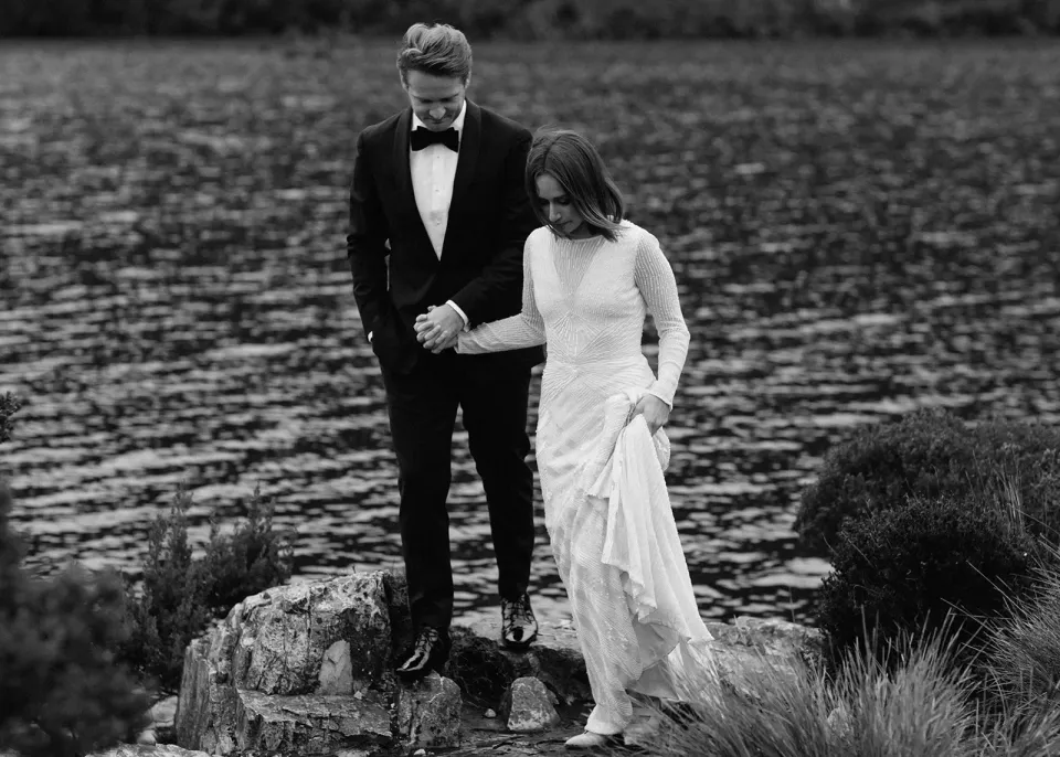 A black-and-white photograph of a couple stepping across rocks beside a lake.