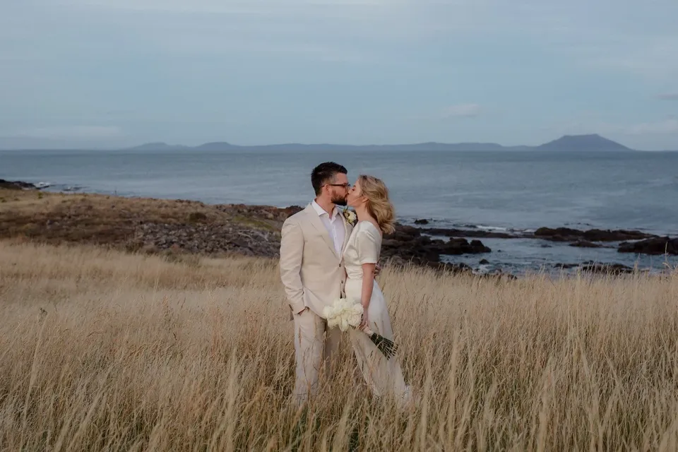 A couple in pale wedding clothes embrace in coastal grass overlooking the sea.