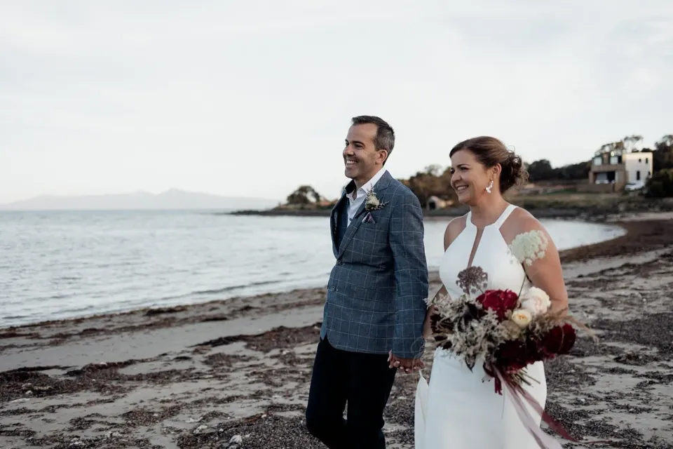 A smiling couple stand on a beach, with the bride holding a red and white bouquet.