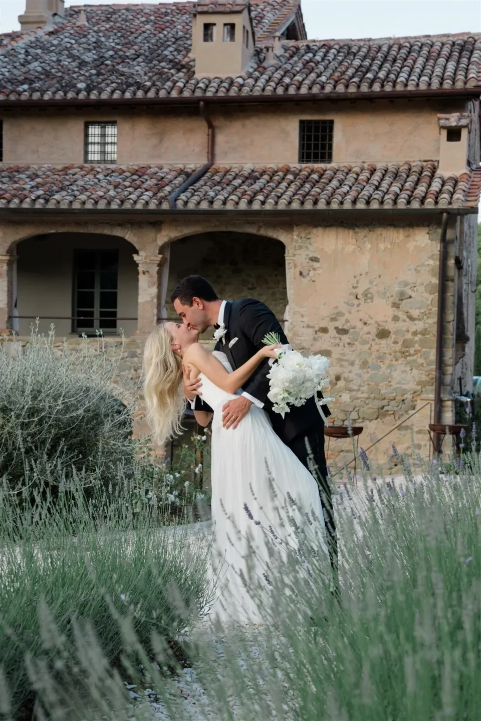 Shelby and Jack kissing among lavender plants beside an old stone building, her bouquet by Le Francesche in hand, in Umbria, Italy, for an Elopement Collective elopement. Photo by James from House of Love.